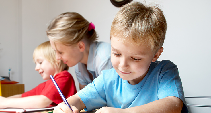 Image of a smiling child, sitting and writing, with a teacher and other child in the background who is doing the same